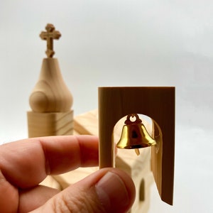 Pode incluir: Um modelo de igreja em miniatura de madeira clara com um sino dourado na torre. O modelo apresenta uma cruz no topo da torre. O sino est&aacute; pendurado em um arco.