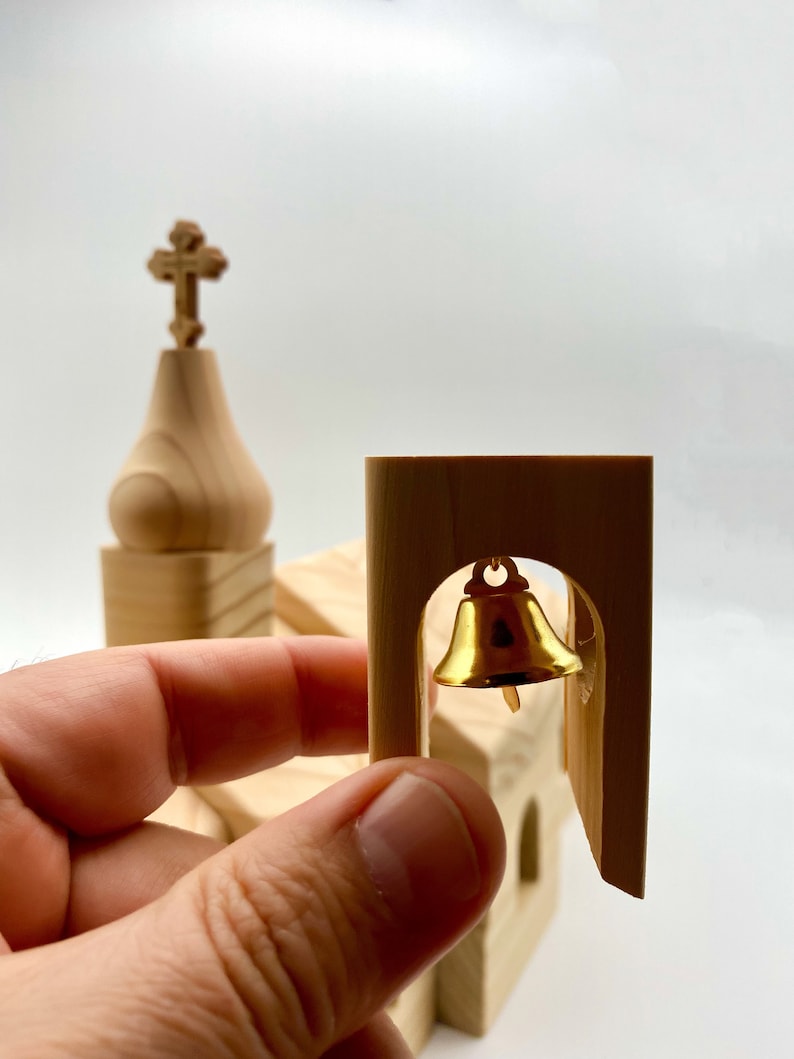 Pode incluir: Um modelo de igreja em miniatura de madeira clara com um sino dourado na torre. O modelo apresenta uma cruz no topo da torre. O sino est&aacute; pendurado em um arco.