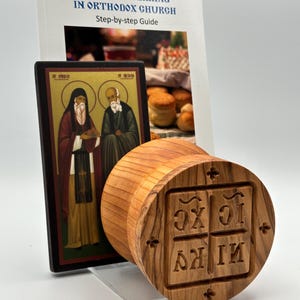 May include: A wooden prosphora stamp with Greek letters and a religious icon, next to a book titled "Prosphora Baking in Orthodox Church." The stamp is cylindrical with a square imprint. The book is a step-by-step guide.