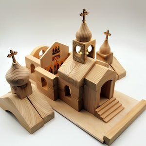 May include: A wooden church building set with multiple pieces. The set includes a main church structure, several smaller buildings, and decorative crosses. The pieces are made of light-colored wood and are arranged on a wooden base.