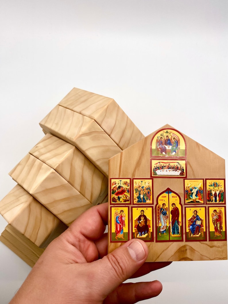 Pode incluir: Um &iacute;cone religioso de madeira com v&aacute;rias imagens pequenas e coloridas que retratam cenas b&iacute;blicas. O &iacute;cone &eacute; segurado em frente a uma pilha de blocos de madeira de cor clara. As imagens s&atilde;o emolduradas em vermelho.