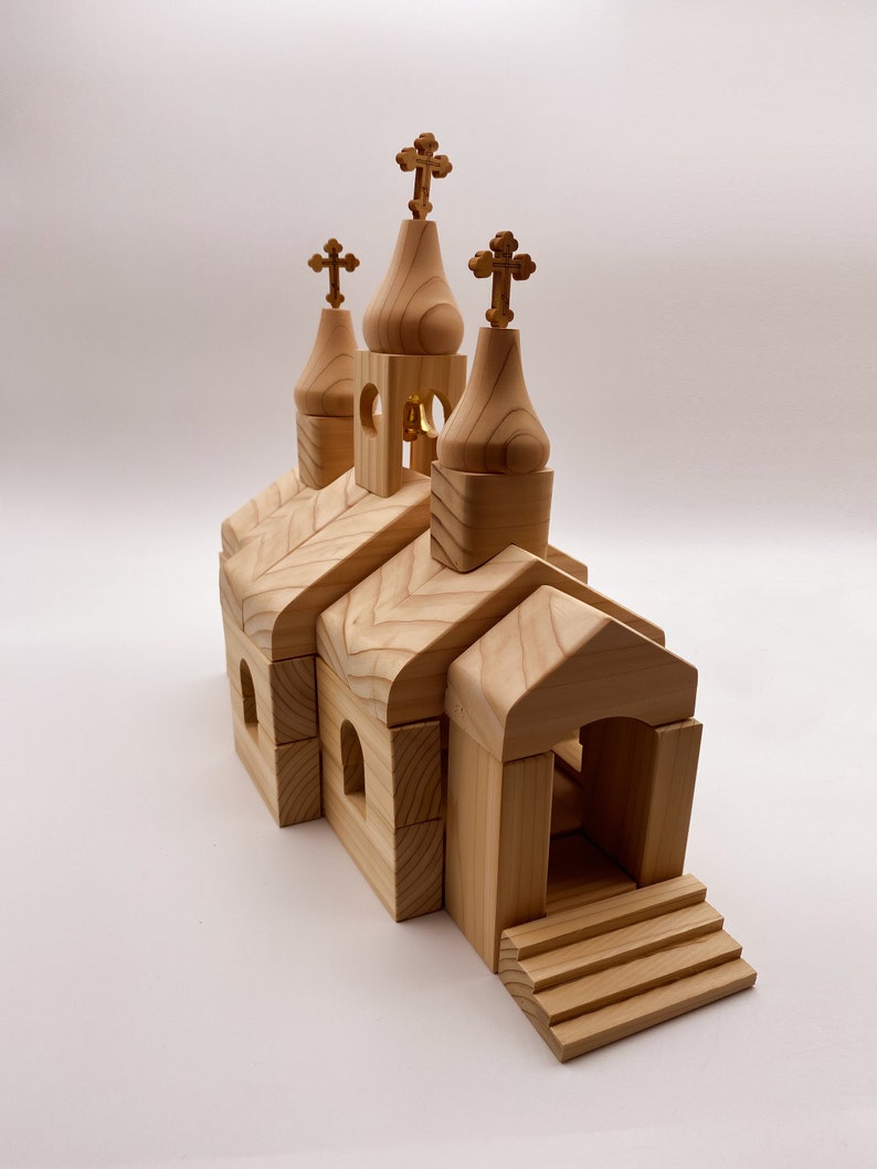 Pode incluir: Maquete de igreja em madeira clara com tr&ecirc;s c&uacute;pulas em forma de cebola encimadas por cruzes. O edif&iacute;cio tem janelas arqueadas e uma entrada com degraus. &Eacute; um brinquedo ou um objeto decorativo.