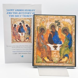 May include: A framed religious icon depicting three figures seated around a table, with halos and robes in shades of blue, orange, and green. The icon is displayed on a clear stand, with a white card behind it that reads "SAINT ANDREI RUBLEV AND THE MYSTERY OF THE HOLY TRINITY."