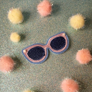 May include: A blue and pink embroidered patch of a pair of sunglasses on a sparkly turquoise background surrounded by small fluffy pom-poms in yellow and pink.