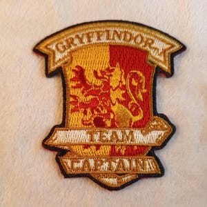 May include: Embroidered Gryffindor crest patch with gold and red colors. The patch features the words "GRYFFINDOR", "TEAM", and "CAPTAIN". The crest includes a lion emblem.