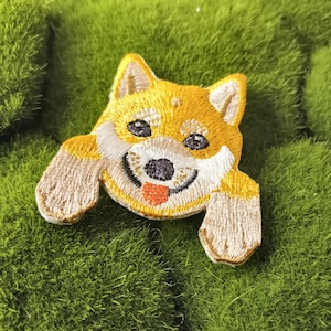 May include: Embroidered patch of a Shiba Inu dog with a happy expression, peeking over the edge of a green surface. The dog is yellow with white markings and a red tongue.