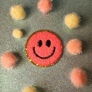 May include: A pink smiley face patch with a black outline and a gold trim surrounded by yellow and pink pom-poms on a sparkly blue background.