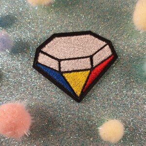 May include: A white diamond-shaped patch with red, yellow, and blue accents. The patch is embroidered with a white diamond shape in the center and a yellow triangle at the bottom.
