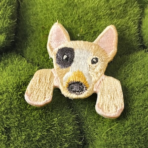 May include: A beige embroidered patch of a dog with black and white markings and pink ears. The dog is looking directly at the camera.