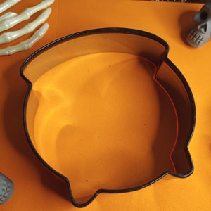 May include: A black cauldron cookie cutter on an orange surface. The cutter is made of metal and has a handle.