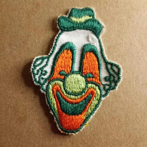 May include: Embroidered clown patch with a smiling face. The patch features a white background with a green hat, orange cheeks, and a green and orange smile. The clown has green hair and is outlined in green.