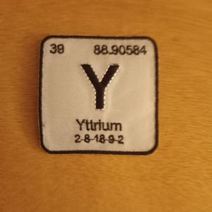 May include: A white rectangular patch with black stitching around the edges. The patch features the chemical symbol "Y" for Yttrium, the atomic number 39, the atomic weight 88.90584, and the electron configuration 2-8-18-9-2.