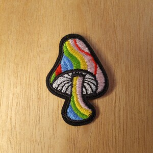 May include: Embroidered mushroom patch with a black border. The mushroom cap features a rainbow design with red, pink, yellow, green, and blue stripes. The stem is white with a pink and yellow stripe.