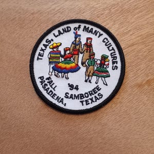 May include: White embroidered patch with a black border. The patch features a circle with the text "Texas, Land of Many Cultures" around the edge. Inside the circle, there are seven children in colorful clothing dancing. The text "Fall '94 Samboree Pasadena Texas" is below the children.