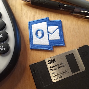 May include: A blue and white embroidered patch with the letter "O" and an envelope, representing the email icon. The patch is on a wooden surface next to a black 3.5-inch floppy disk labeled "High Density Haute densité IBM Formatted 1.44 MB".