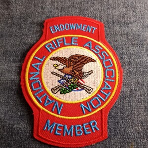 May include: Red, white, and blue embroidered patch with the text "National Rifle Association Endowment Member". A bald eagle with crossed rifles is in the center of the patch.
