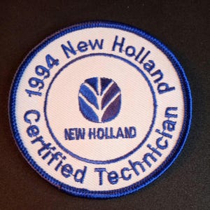 May include: A circular patch with a white background and blue embroidered text and accents. The text reads "1994 New Holland Certified Technician" around the perimeter. The center features a blue logo and the words "NEW HOLLAND".