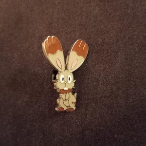 May include: A metal pin featuring a cartoon rabbit with large ears. The rabbit has a tan body, white face, and brown accents on its ears. The pin is set against a dark brown background.
