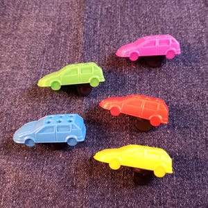 May include: Five colorful toy cars in pink, green, red, blue, and yellow are arranged on a denim background. Each car is a different color and has a black base. The cars are small and appear to be made of plastic.