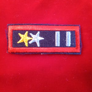May include: A rectangular patch with a navy blue background, framed by a red border. The patch features a yellow star, a white star, and two light blue vertical bars. The patch is set against a red background.