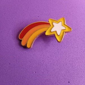 May include: A gold-colored enamel pin with a rainbow shooting star design. The rainbow is red, orange, yellow, and gold. The star is yellow with a white center.