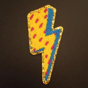 May include: A yellow lightning bolt with pink and blue accents. The bolt is outlined in gold glitter.
