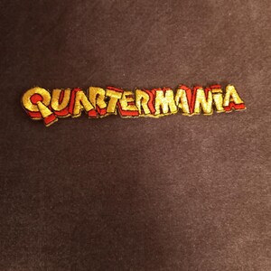 May include: A gold and red embroidered patch with the word "Quartermania" in a bold, stylized font.