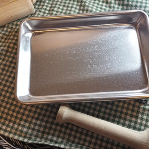 May include: A silver baking sheet with a slightly textured surface sits on a green and white checkered tablecloth. A wooden rolling pin is also visible in the image.