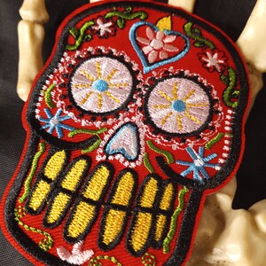 May include: Red embroidered sugar skull patch with yellow teeth, white eyes with blue centers, and floral details.
