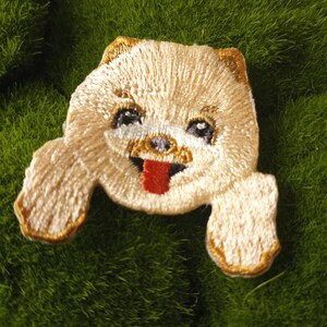 May include: Embroidered patch of a white dog with a red tongue sticking out. The dog is looking up with its eyes closed. The patch is on a green background.