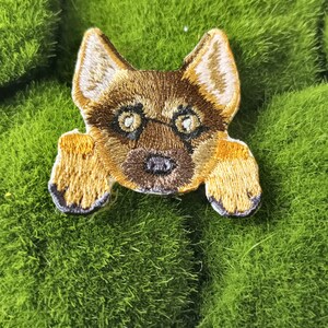May include: A brown and tan embroidered patch of a German Shepherd dog peeking over a green surface. The dog has brown eyes and a black nose.