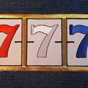May include: Embroidered patch featuring three slot machine reels, each displaying a number seven in red, white, and blue. The patch has a gold border and is set against a denim background.