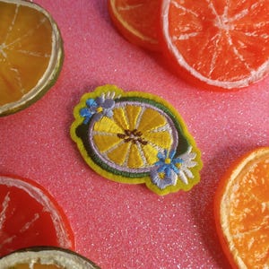 May include: A yellow embroidered patch in the shape of a lemon slice, with blue flower accents, sits on a pink, glittery surface. Orange and yellow citrus slices are scattered around the patch, creating a vibrant, fruity theme.