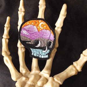 May include: Black embroidered skull patch with a purple, orange, and blue mountain scene inside. The scene includes a crescent moon and stars.