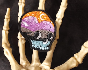 May include: Black embroidered skull patch with a purple, orange, and blue mountain scene inside. The scene includes a crescent moon and stars.