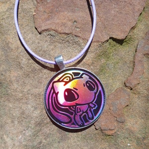 May include: A round pendant necklace with a cartoon character design. The pendant has a silver-colored border and a holographic image with pink, purple, and yellow hues. The necklace cord is a light purple color.
