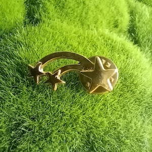 May include: Gold-toned shooting star pin with three stars and a round base. The pin is on a green background.