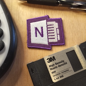 May include: Purple and white embroidered patch with the letter "N" and a series of lines. The patch is attached to a 3M High Density Haute densité IBM Formatted - Formattée 1.44 MB floppy disk.