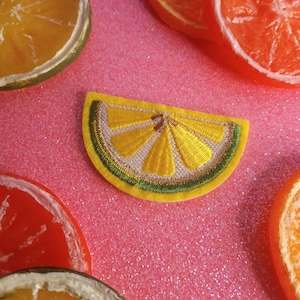 May include: A yellow embroidered lemon slice patch with green stitching on a pink sparkly background.