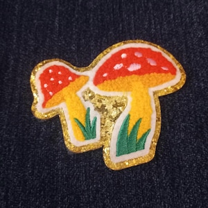 May include: A colorful embroidered patch featuring two mushrooms. The mushrooms have red caps with white spots, yellow stems, and green grass. The patch is outlined in gold glitter and white. The background is a dark blue.