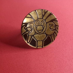 May include: A gold-colored metal pin with a black background. The pin features three cartoon faces with different expressions. The faces are surrounded by a decorative design.