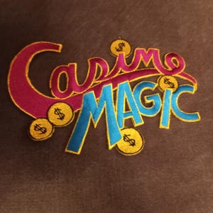 May include: Embroidered patch with the words "Casino MAGIC" in a stylized font. The text is in blue and pink with gold trim, and gold coin accents.