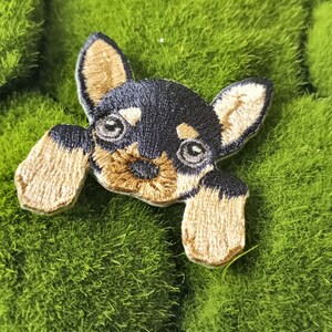 May include: A black and brown embroidered patch of a dog peeking over a green surface. The dog has brown ears and a black nose.