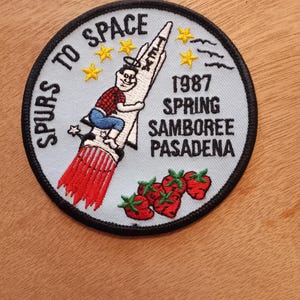 May include: A white and blue embroidered patch with a cartoon character riding a rocket. The text "SPURS TO SPACE" is written in a circle around the rocket. The text "1987 SPRING SAMBOREE PASADENA" is written below the rocket. There are five yellow stars and three red strawberries on the patch.