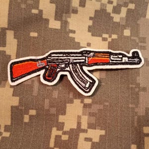 May include: Embroidered patch featuring a stylized AK-47 assault rifle. The patch has a white border, with the rifle depicted in black, silver, and orange. The background is a camouflage pattern.