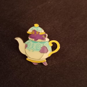 May include: A purple and yellow teapot pin with a whimsical design. The teapot has a blue and white swirl pattern and a yellow lid. A purple frog is peeking out of the teapot.
