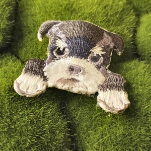 May include: A small, black and white embroidered patch of a miniature schnauzer dog with white fur on its paws and chest. The patch is on a green mossy surface.