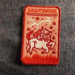 May include: Red embroidered patch with the word "Sagittarius" and a white illustration of a centaur with a bow and arrow.