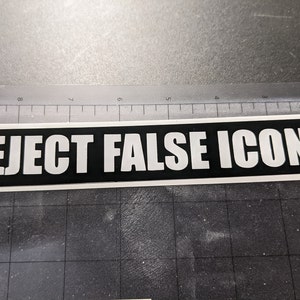 May include: A rectangular sticker with the words "REJECT FALSE ICONS" in bold white capital letters on a black background. The sticker is on a gray surface with a ruler in the background.