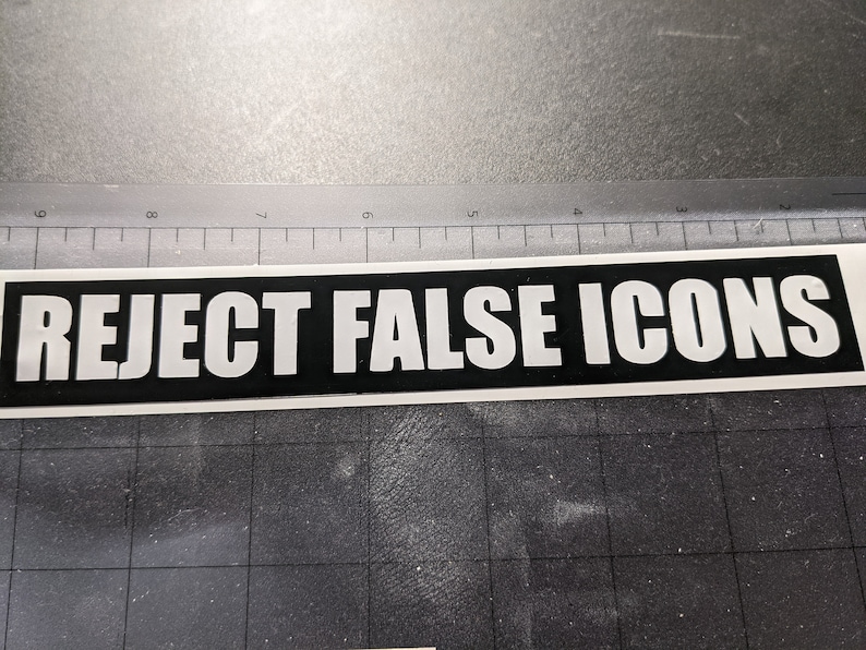 May include: A rectangular sticker with the words "REJECT FALSE ICONS" in bold white capital letters on a black background. The sticker is on a grey surface with a ruler in the background.